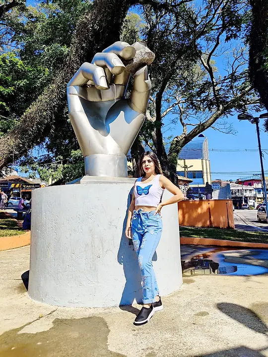 Modelo de cropped branco e calça jeans ao lado de uma escultura de mão numa praça