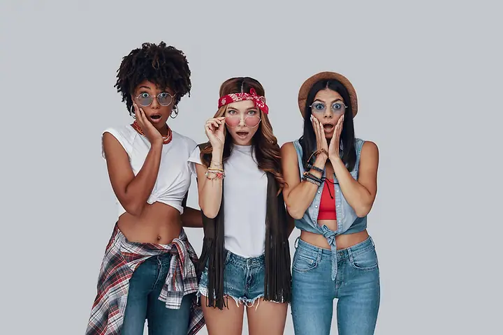 Três mulheres de calça jeans e camiseta, fazendo cara de surpresa